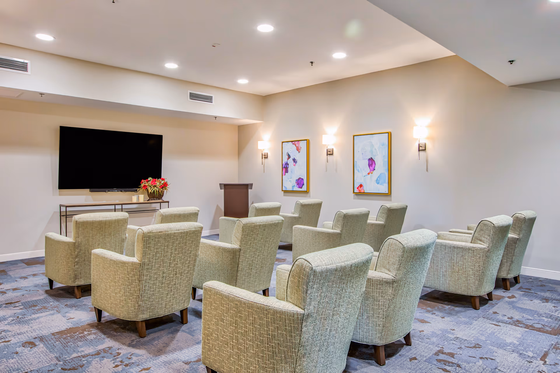 A small presentation or media room with two rows of light green upholstered armchairs facing a large flat-screen TV mounted on the wall. There is a podium in front of the chairs and two abstract paintings on the wall with wall-mounted lights above them. The room has a patterned carpet and recessed ceiling lights.