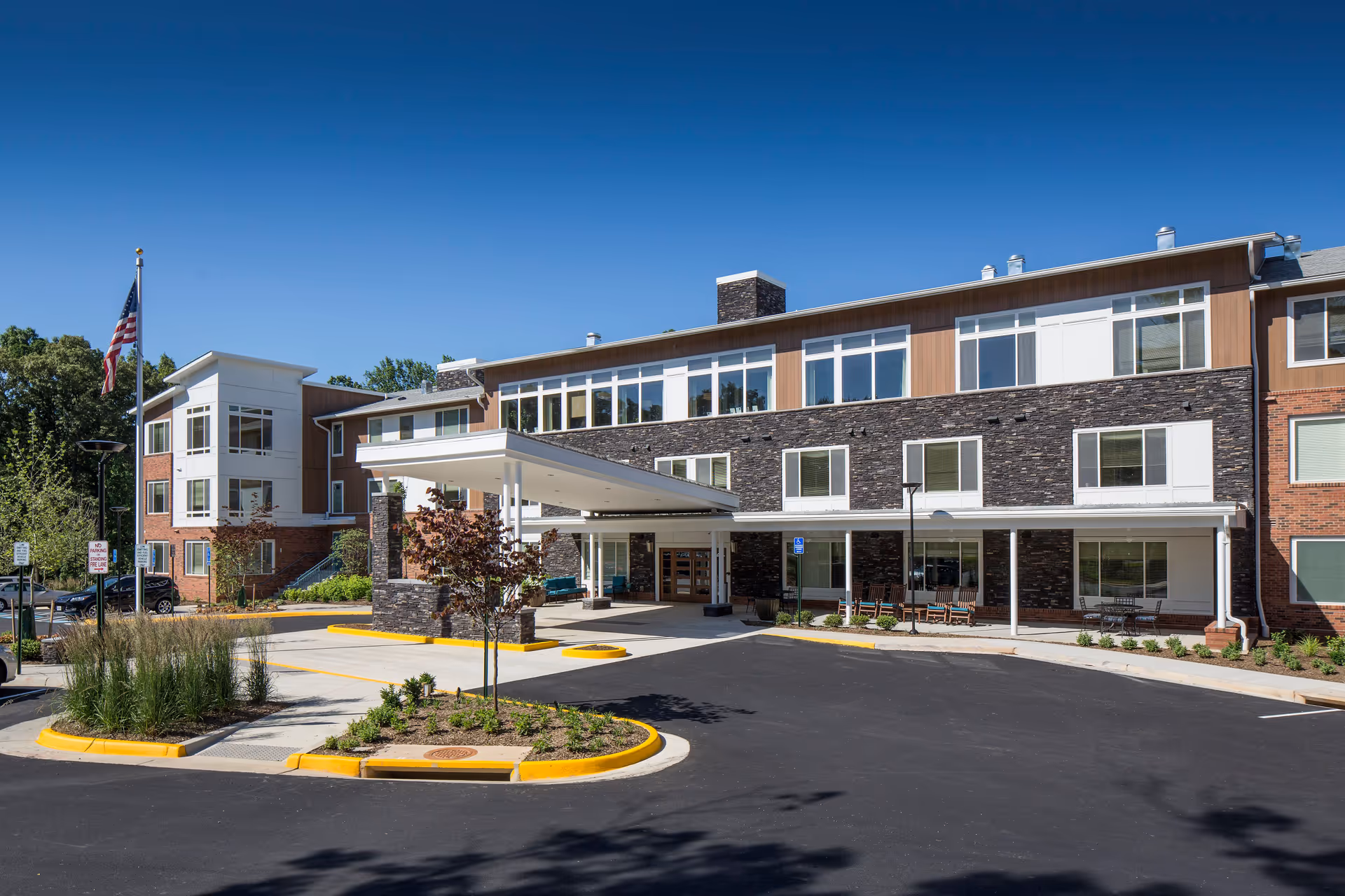 Front entrance of a three-story senior living facility with a covered drop-off, landscaped islands, and an American flag.