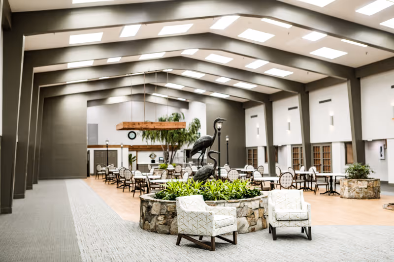 Spacious senior living common area with dining tables, lounge chairs, a central stone planter with bird sculptures, and a high beamed ceiling with skylights.