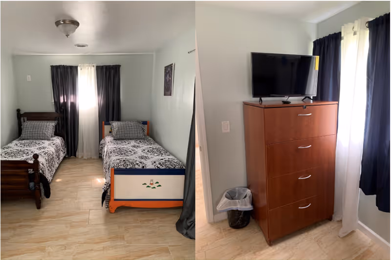 A bedroom with two single beds, each with patterned bedding and pillows, positioned side by side against a light-colored wall with a window covered by dark curtains. Adjacent to the beds is a wooden dresser with four drawers, a flat-screen TV on top, and a small trash can beside it. The room has light-colored tiled flooring and light walls.