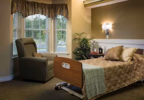 Senior living bedroom featuring a bed, armchair, windows with curtains, and ambient lighting.