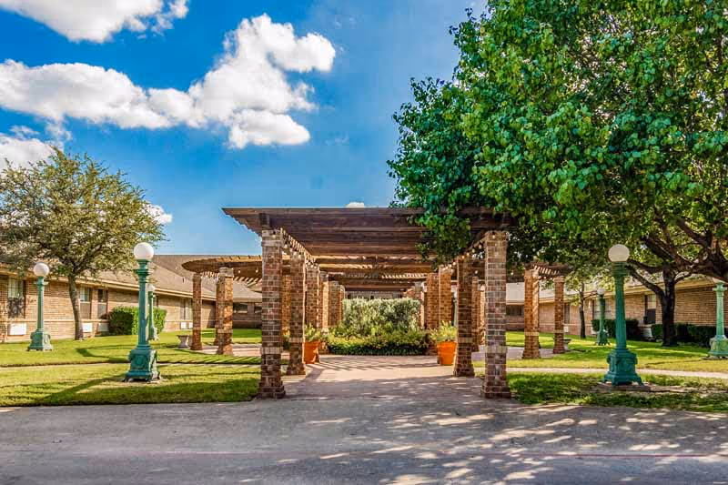 Brick pergola-covered walkway through a green courtyard with trees and vintage lampposts in front of a single-story senior living building.
