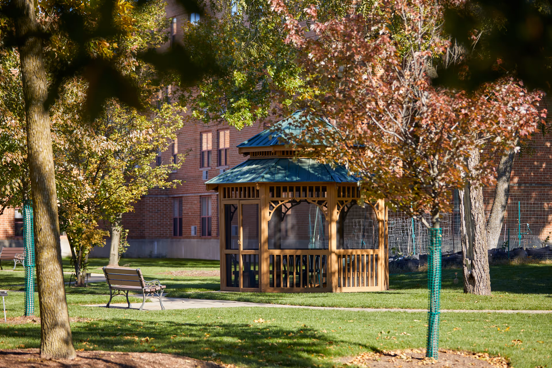 A wooden gazebo in a grassy courtyard surrounded by trees and benches with a brick building in the background.