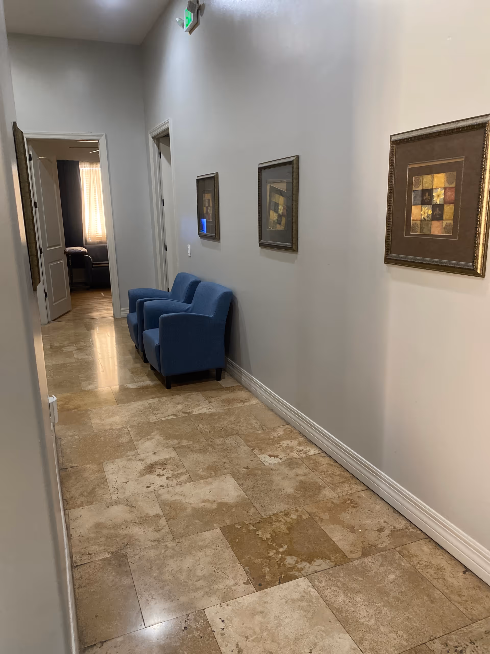 Hallway with two blue armchairs along a beige tiled floor, framed pictures on the wall, and an open doorway at the far end.