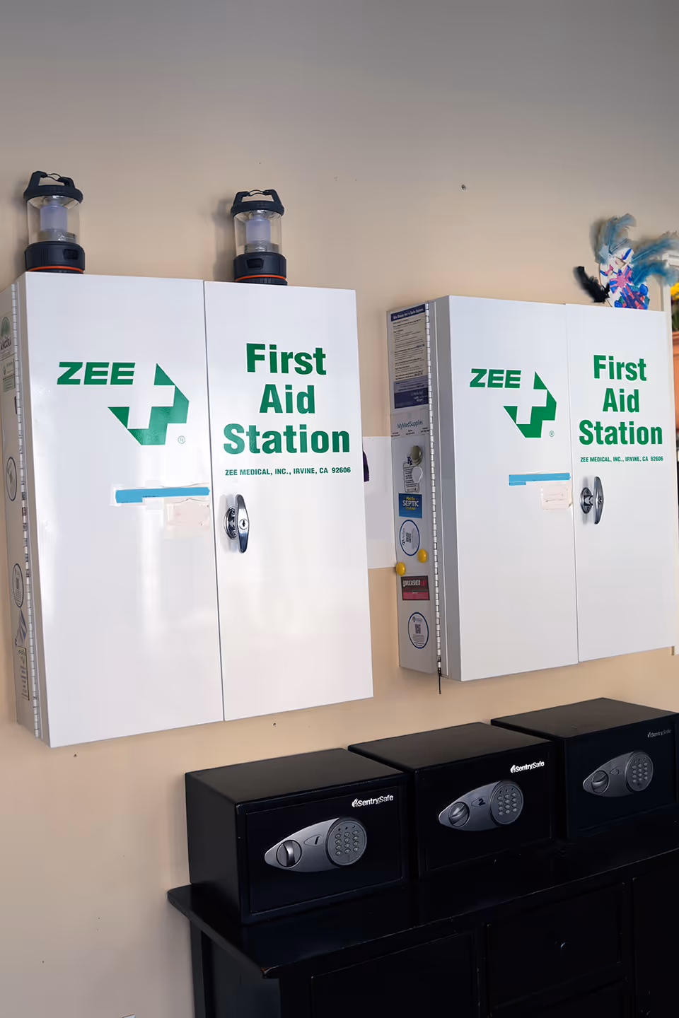 Wall-mounted white First Aid Station cabinets with black safes beneath in a hallway.