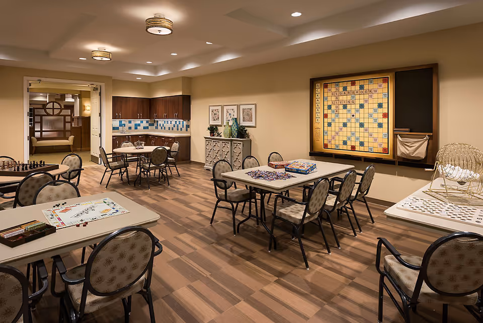 A well-lit activity room with multiple tables and chairs arranged for games and socializing. The tables have board games like Monopoly, Scrabble, and puzzles set up. The walls are beige with a large Scrabble board mounted on one side. There is a small kitchenette area with cabinets and a tiled backsplash in the background. The room has a warm and inviting atmosphere with wood-patterned flooring and ceiling lights.
