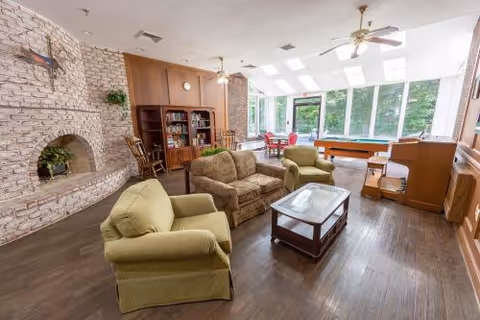 Bright communal living room with sofas and armchairs around a coffee table, a brick fireplace and bookshelves, a pool table, and large windows overlooking greenery.