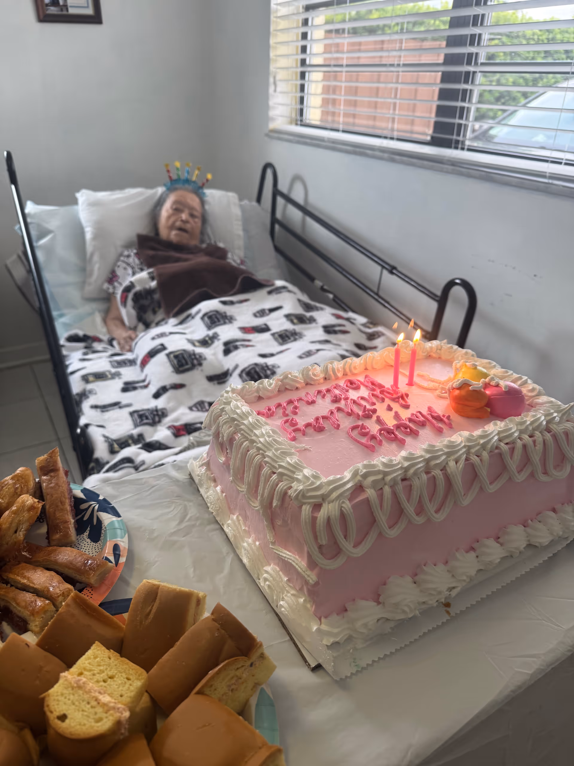 An elderly resident lies in a bed beside a pink birthday cake on a table.