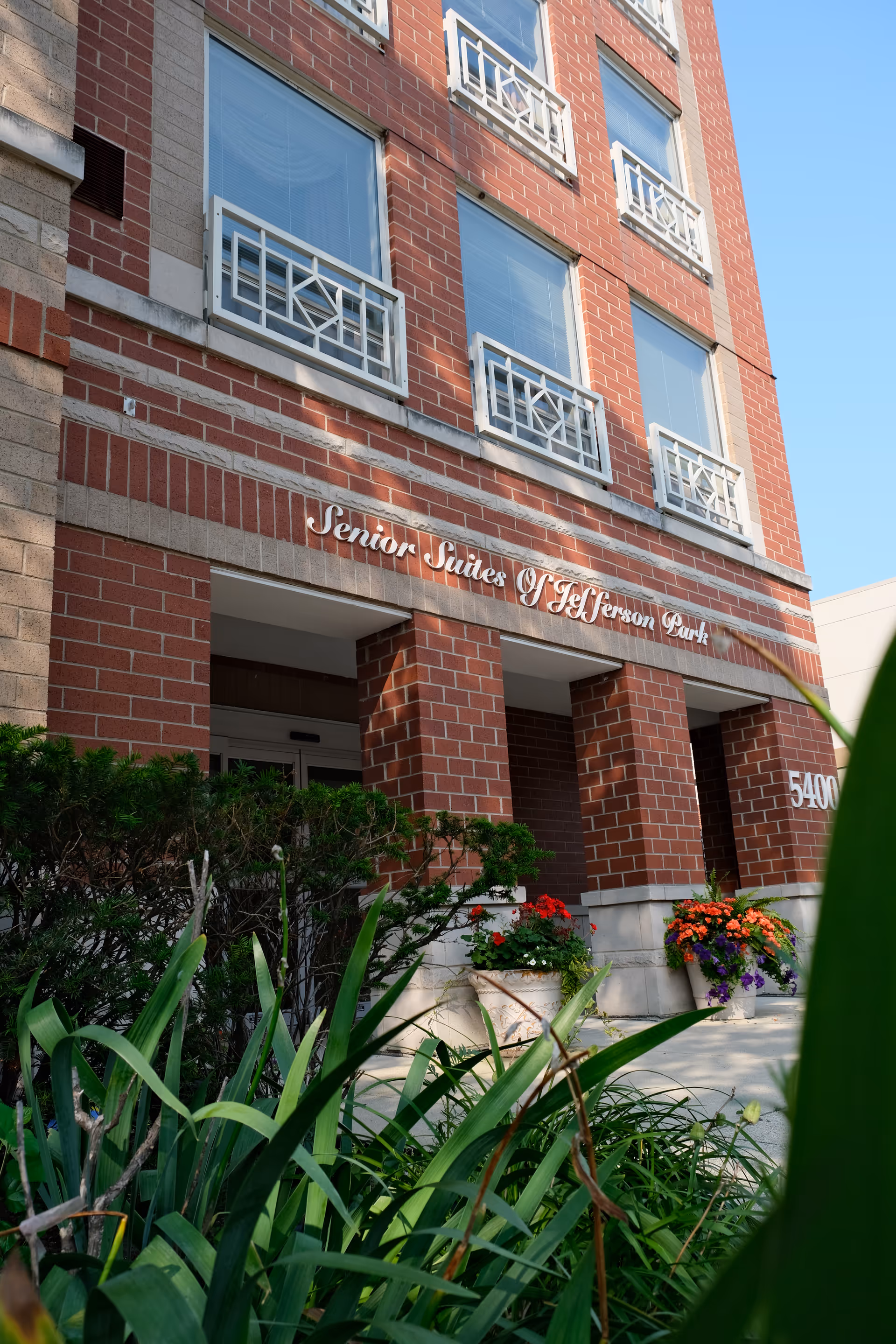 Entrance of a red-brick building labeled 'Senior Suites of Jefferson Park' with windows above, planters of flowers and greenery in front.