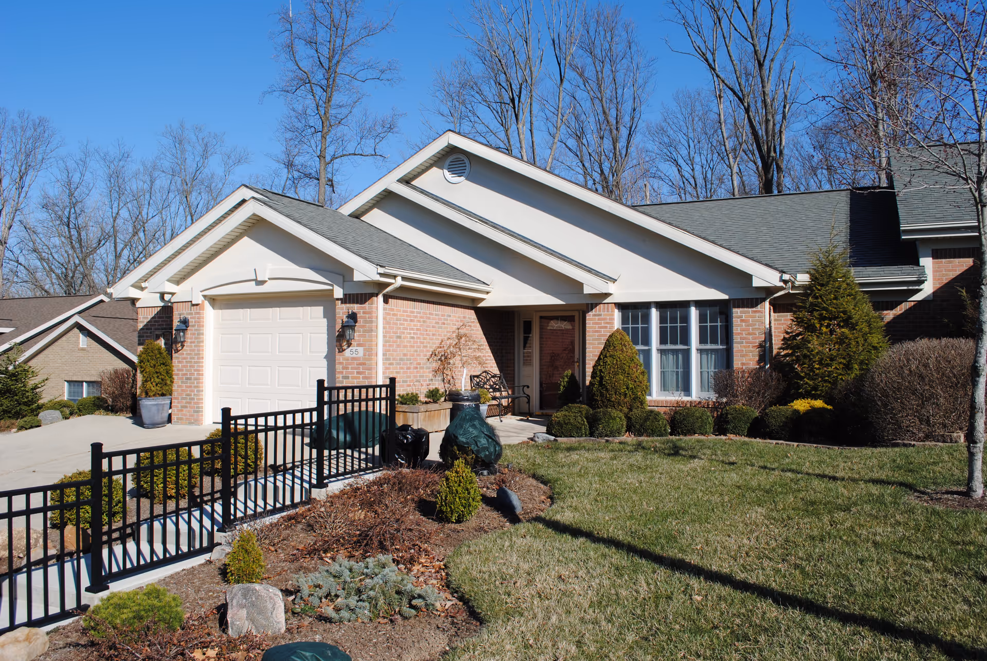Exterior view of a single-story brick house with a garage, a front door, and a well-maintained lawn and garden. There are leafless trees in the background under a clear blue sky.