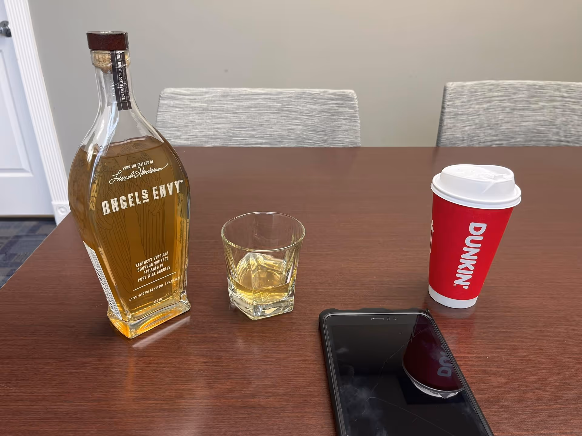 A bottle of Angels Envy whiskey, a glass, a Dunkin cup, and a smartphone on table.
