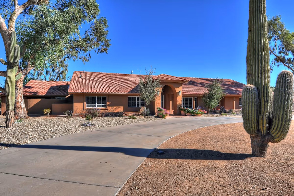 Single-story stucco building with a red tile roof, desert landscaping and large saguaro cacti beside a curved driveway under a clear blue sky.