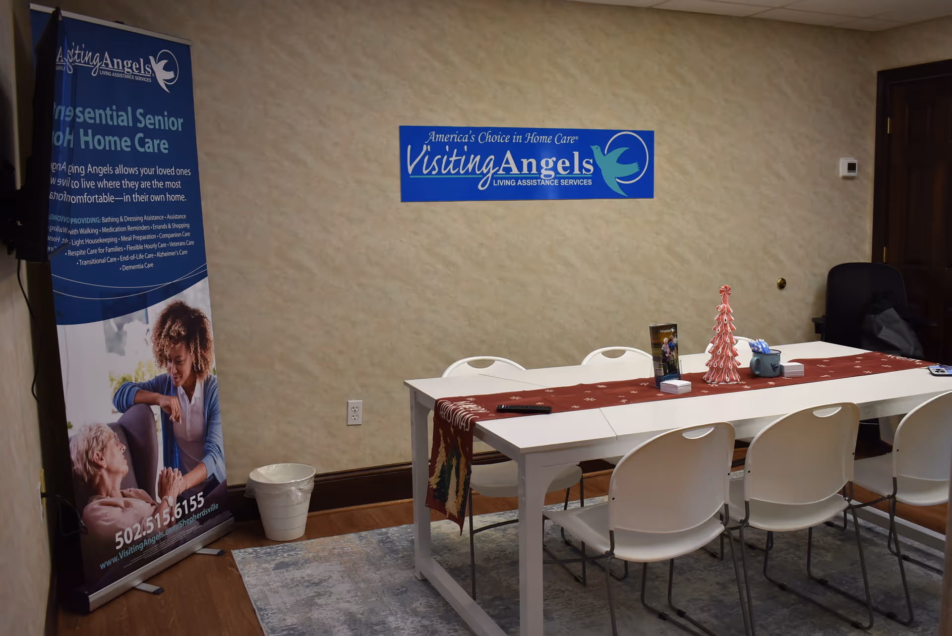 Conference table with white chairs in a beige-walled room featuring Visiting Angels signage.