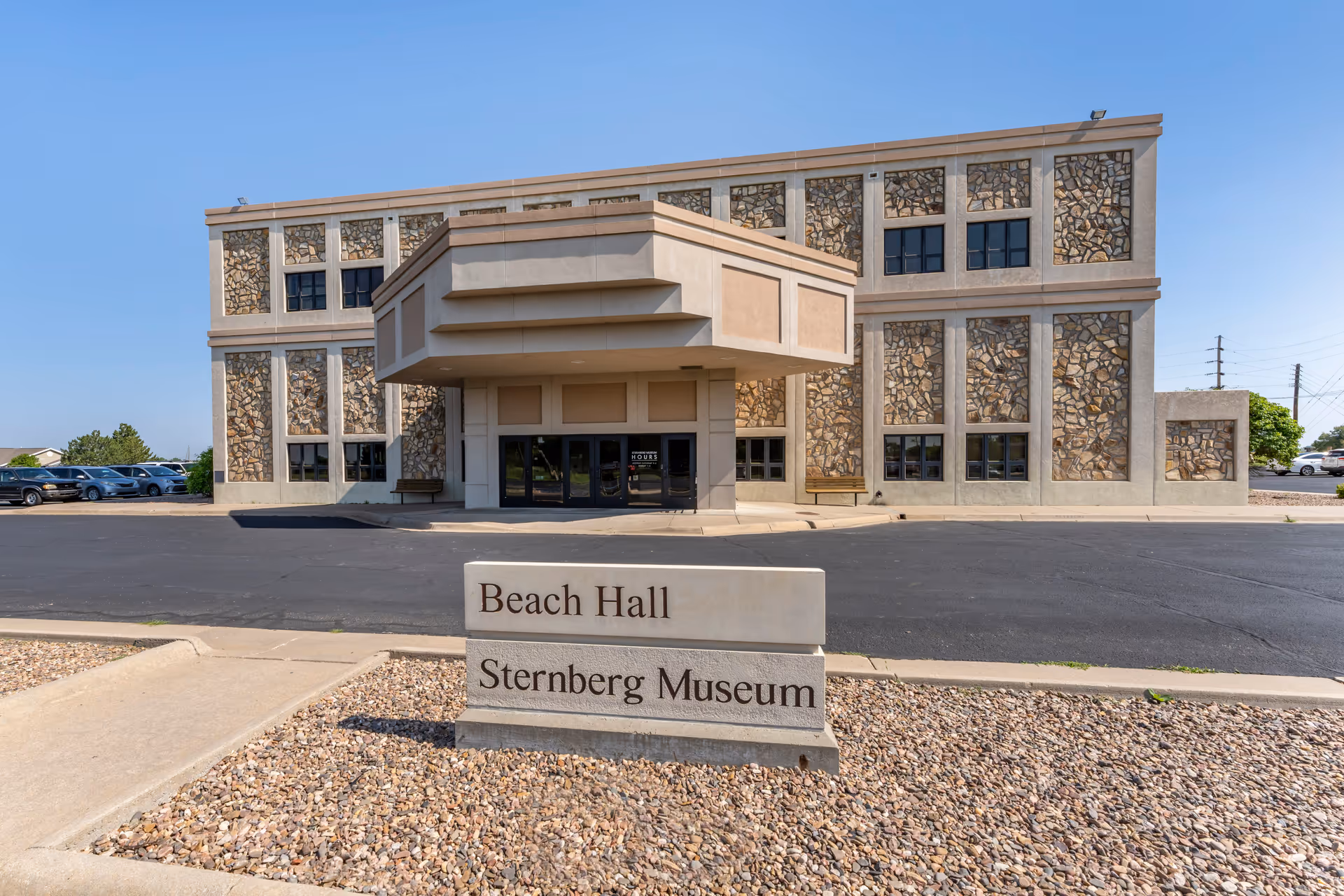 Exterior view of a three-story building with stone and concrete facade under a clear blue sky. In front of the building, there is a sign that reads 'Beach Hall Sternberg Museum'. Several parked cars are visible to the left side of the building.