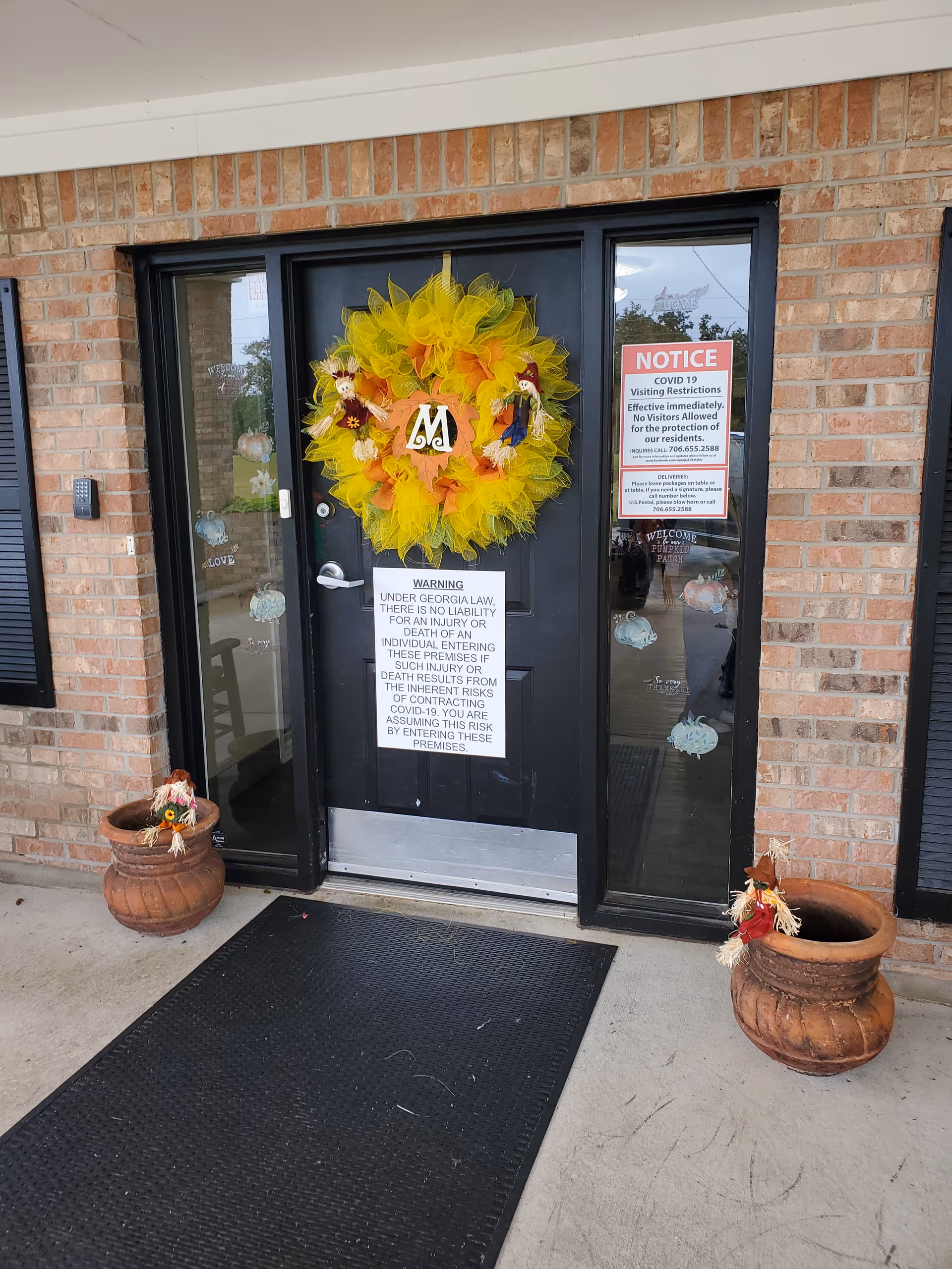 Entrance door of a facility decorated with a large yellow and orange wreath featuring small scarecrow figures. The door is black with glass panels on either side. There are two large terracotta pots on either side of the door, each with a small scarecrow decoration. Several signs are posted on the door, including a COVID-19 visiting restrictions notice and a legal warning about liability under Georgia law. The building exterior is brick with black shutters and a black rubber mat in front of the door.