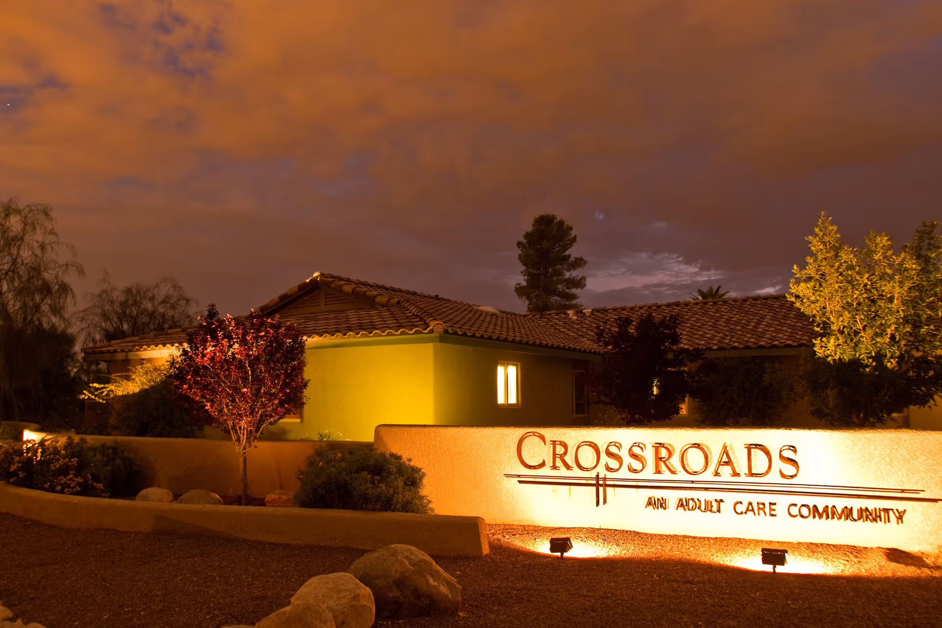 Nighttime view of the illuminated front of the Crossroads assisted living building and its lit sign amid landscaped grounds.