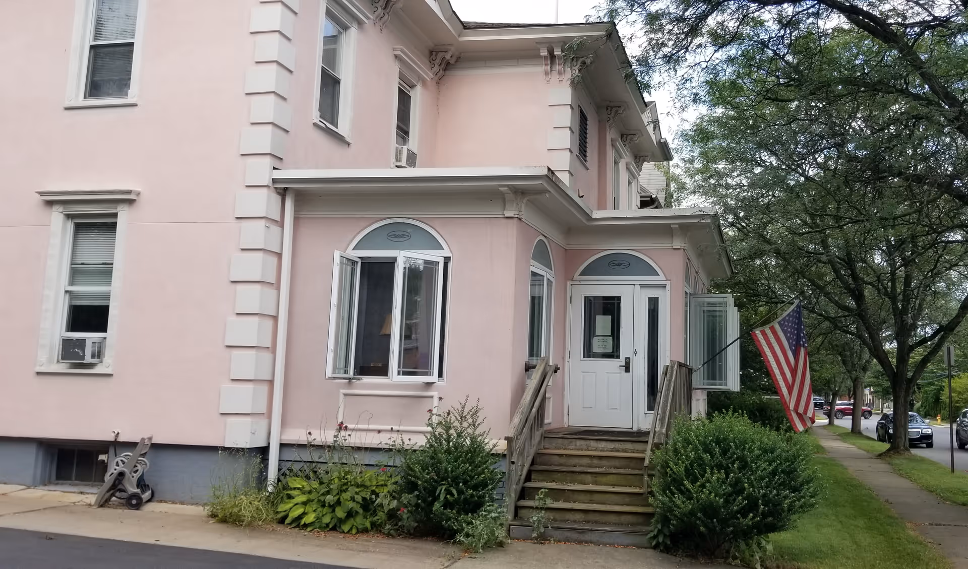 Pink two-story senior care home with front entrance, stairs, and American flag.