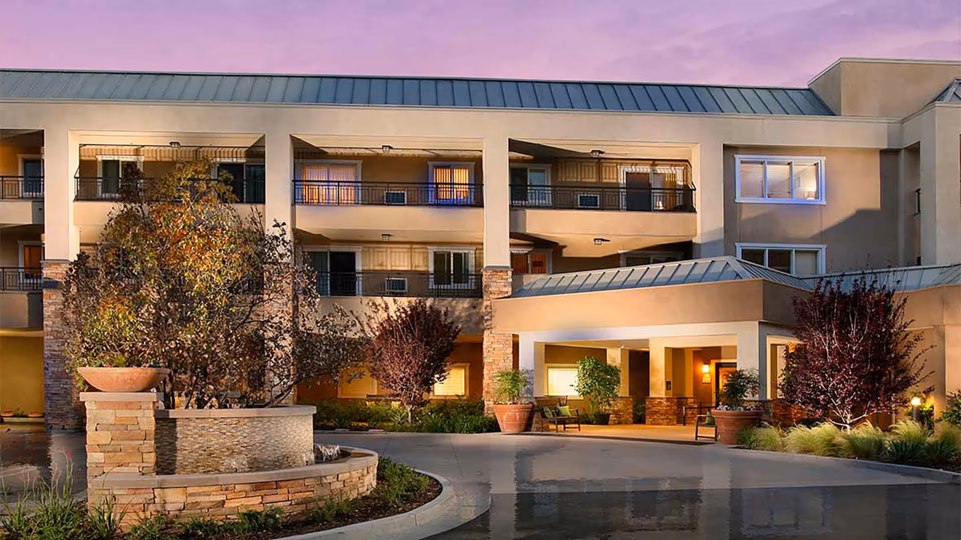Exterior view of a multi-story senior living facility building at dusk with a covered entrance, balconies, and landscaped area including trees and a stone planter with a water feature.