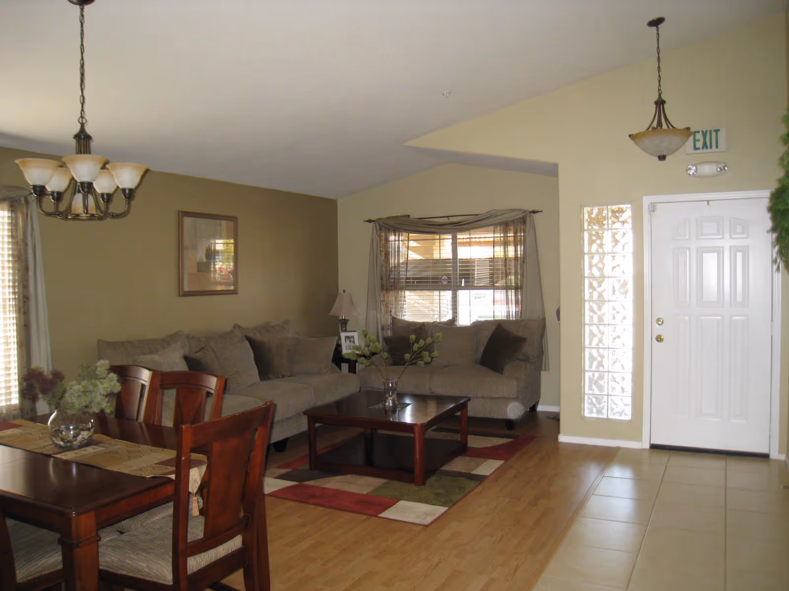 Interior view of a living room and dining area in an assisted living facility. The living room has two beige sofas with cushions, a wooden coffee table on a multicolored rug, and a window with sheer curtains. The dining area features a wooden dining table with four chairs and a floral centerpiece. The floor is a combination of light wood and beige tiles. There is a white door with an exit sign above it and a hanging light fixture near the door.