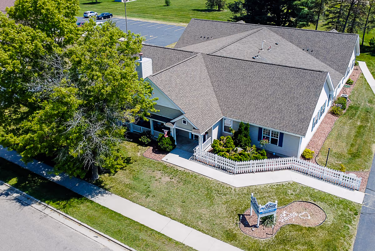 Aerial view of a single-story senior living memory care building with a white picket fence, entrance porch, landscaped lawn and a sign near the sidewalk.