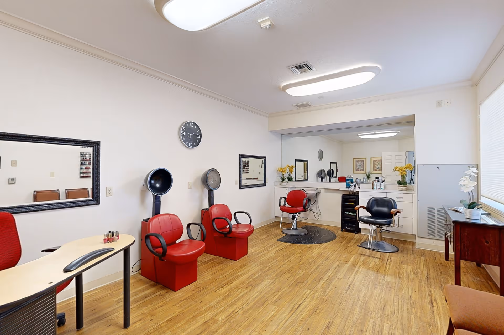 Interior view of a salon area with wooden flooring, featuring two red salon chairs with hair dryers, two styling chairs in front of a large mirror, a small table with nail polish, and a window with blinds letting in natural light.