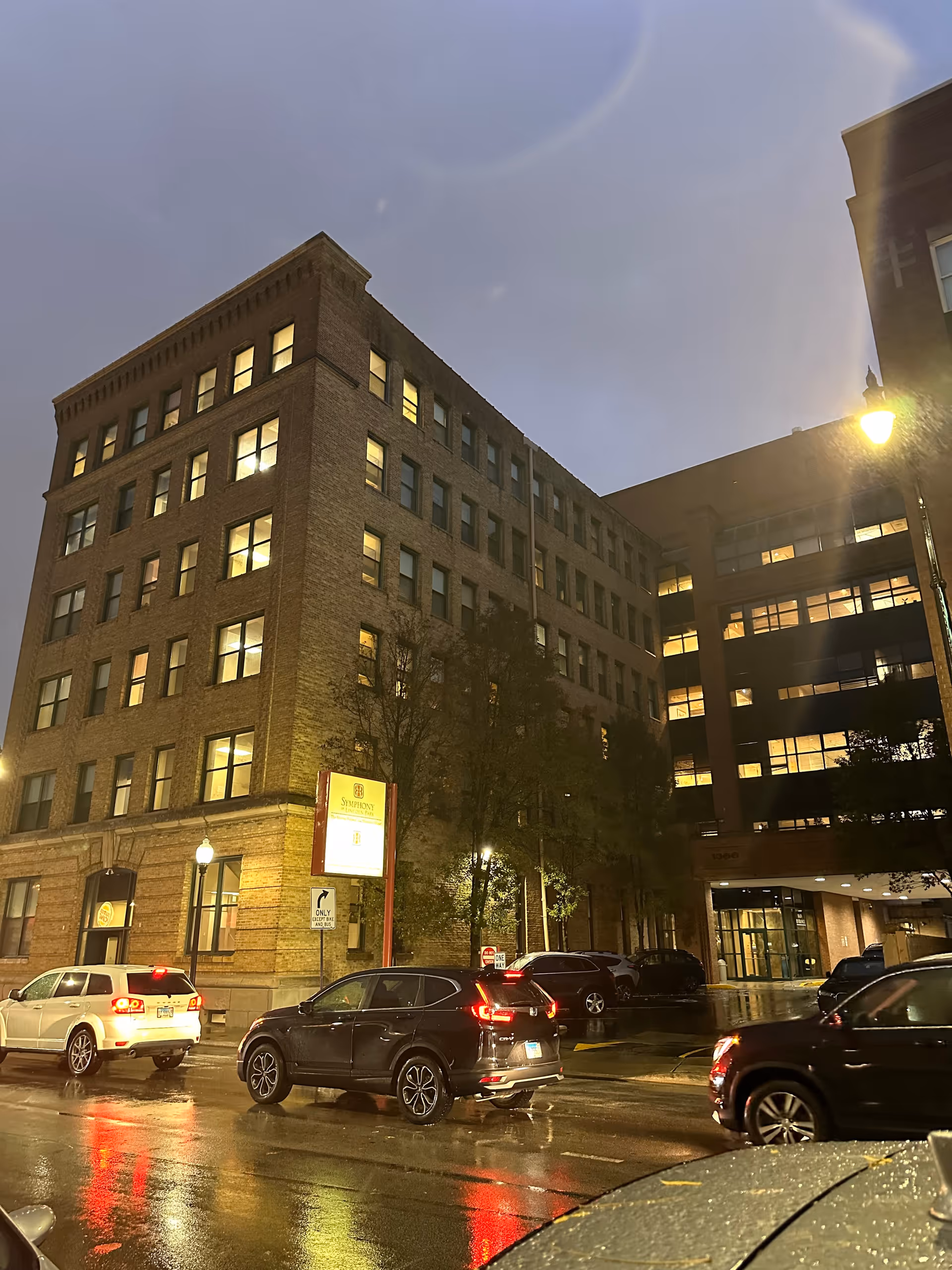 A nighttime street view of a multi-story brick building with many lit windows. Several cars are parked and driving on the wet street in front of the building. There is a street lamp illuminating the scene and a signpost near the building entrance.
