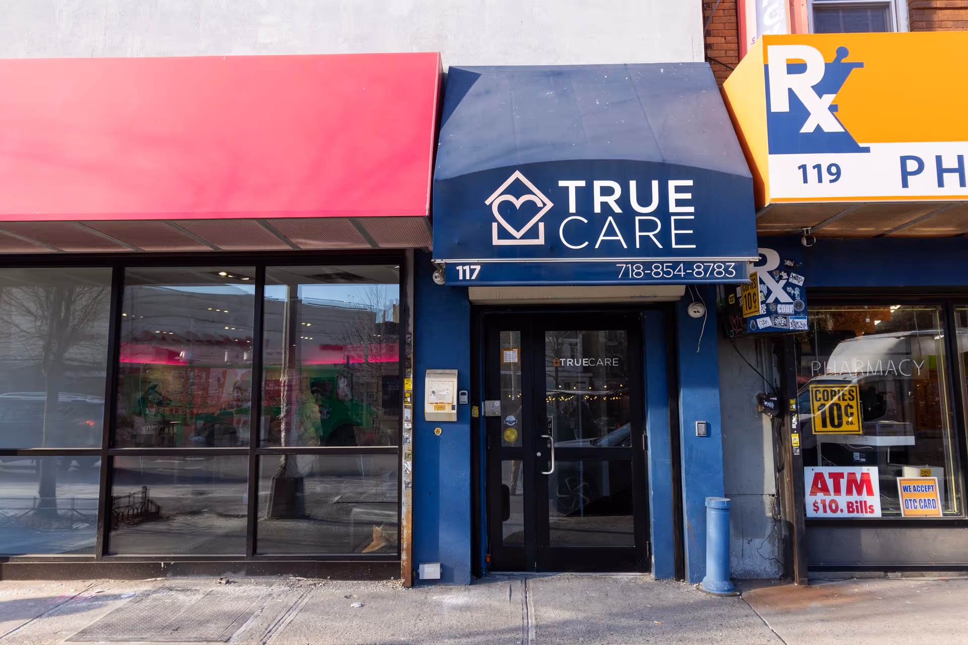 Blue storefront with True Care signage and glass doors, adjacent pharmacy storefront, on sidewalk.