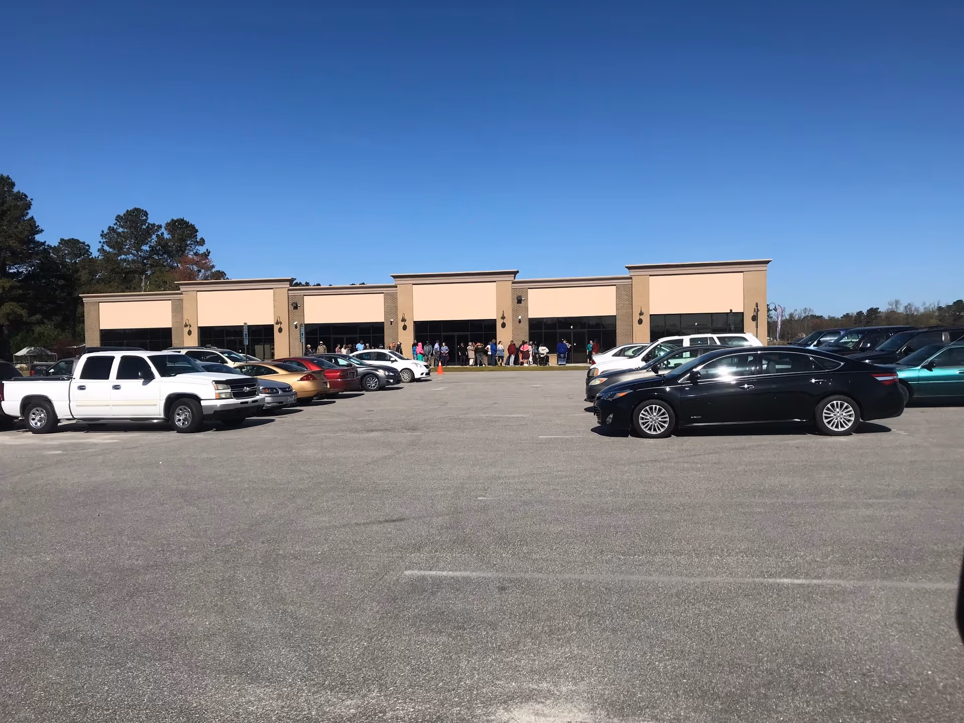 Parking lot in front of a beige, single-story building with people near the entrance.