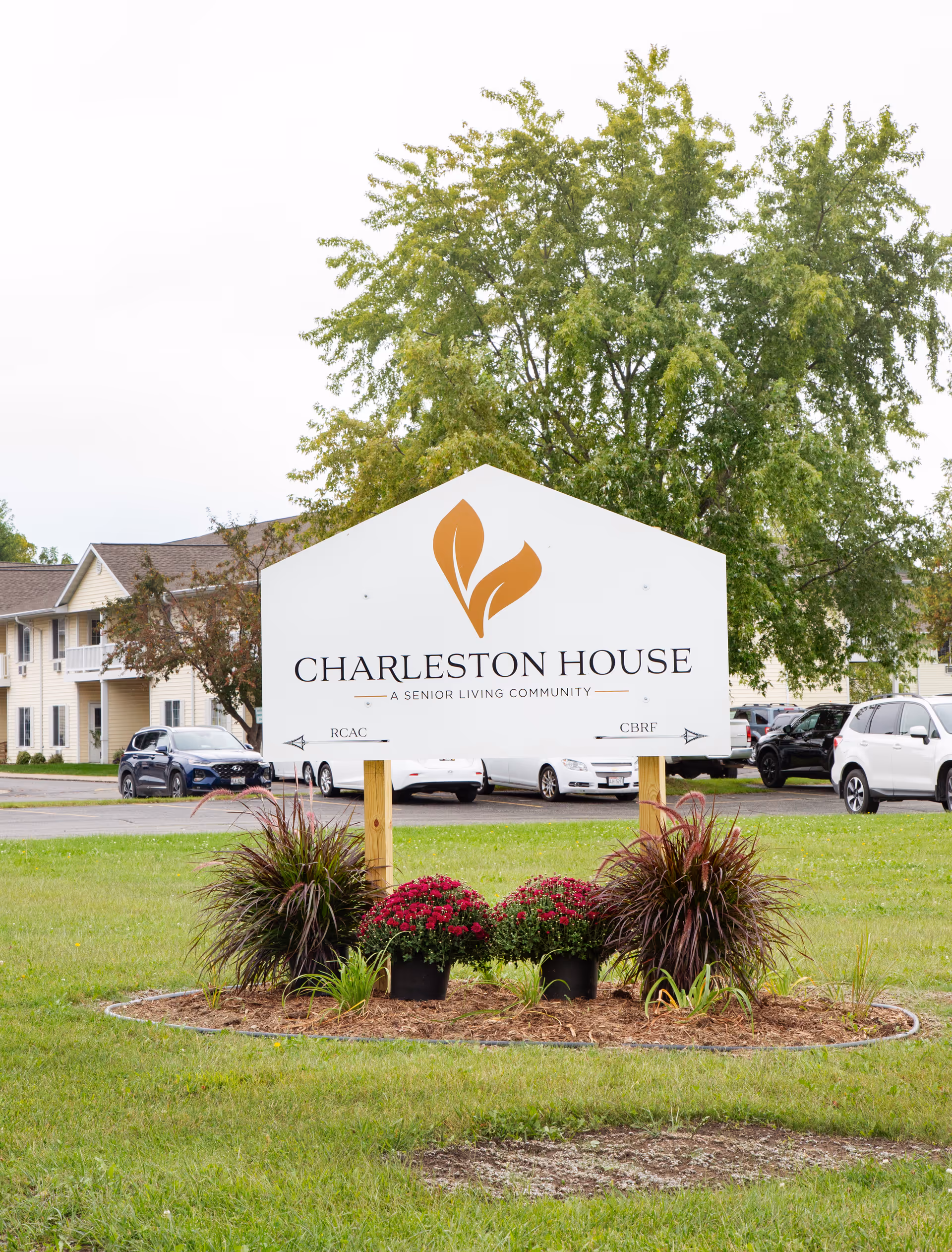 White Charleston House Assisted Living sign on a grassy lawn with flowers, parked cars, and the facility building behind it.