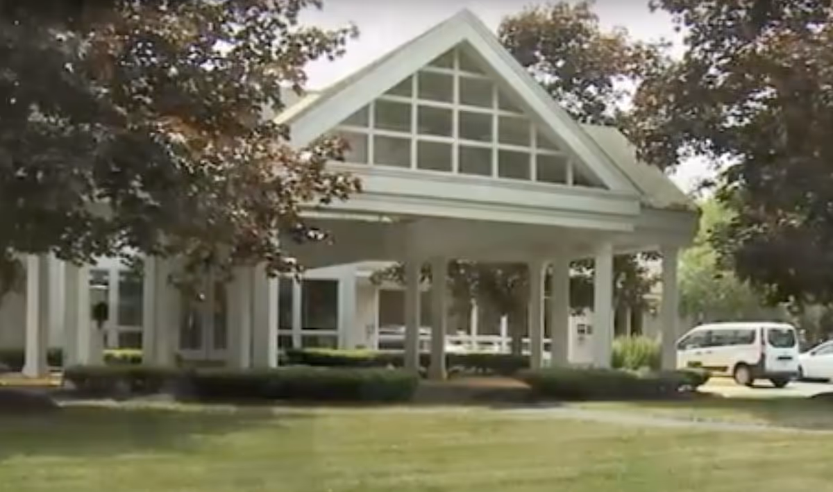 White senior living facility entrance with a porte-cochère, columns, and parked cars.