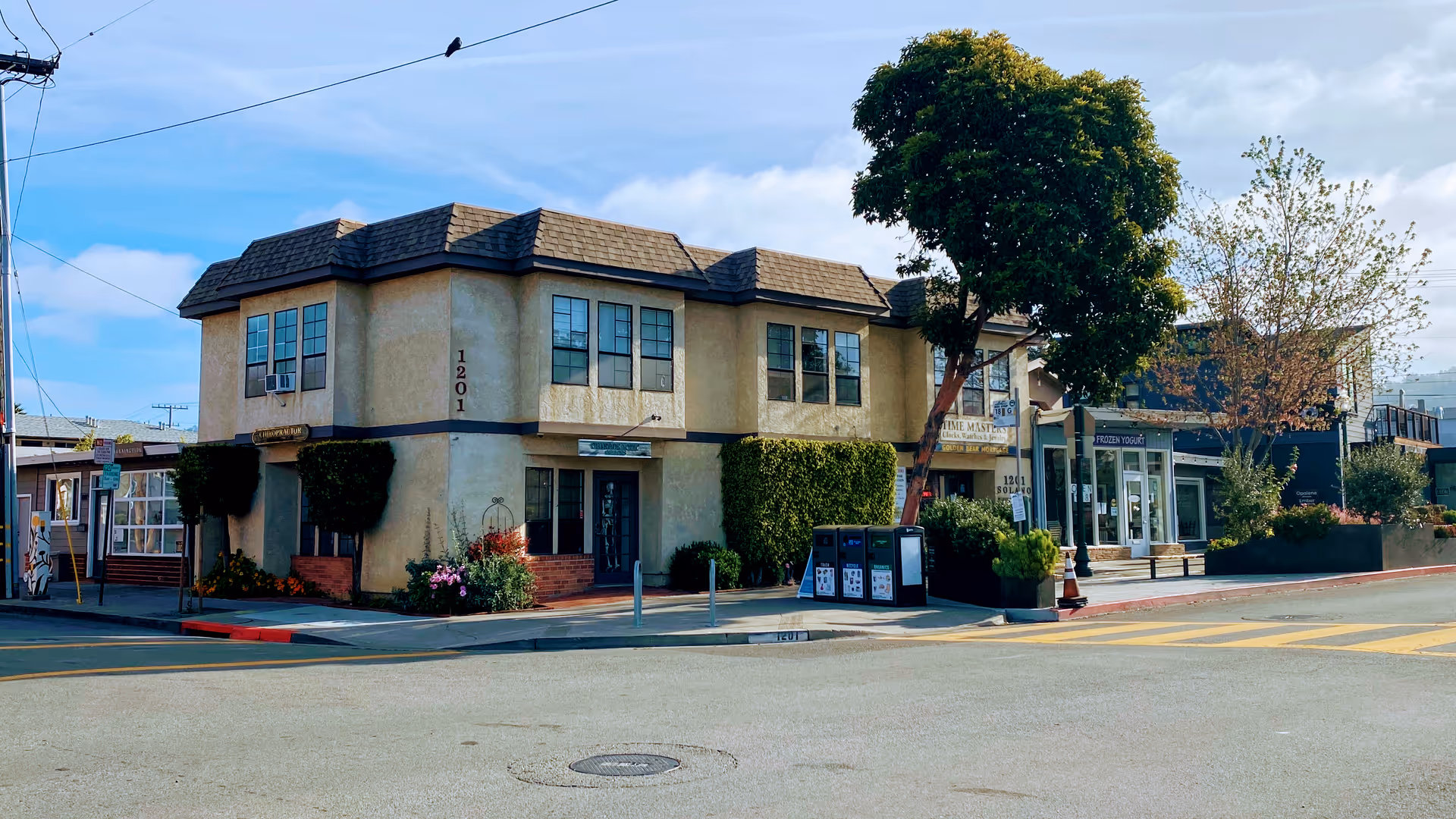 Two-story beige commercial building on a sunny street with storefronts and trees