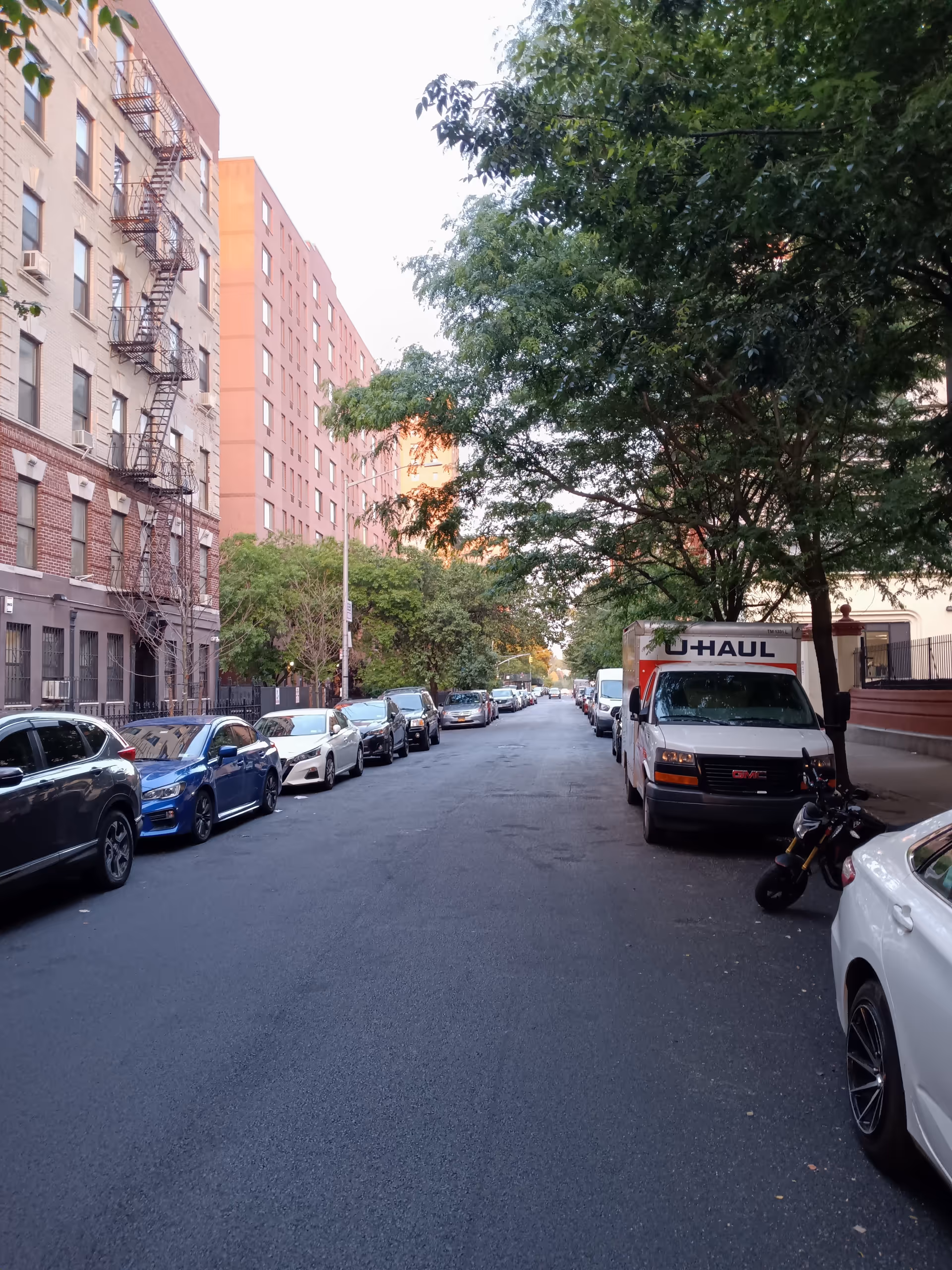 City street with parked cars, a U-Haul truck, and apartment buildings lined by trees.