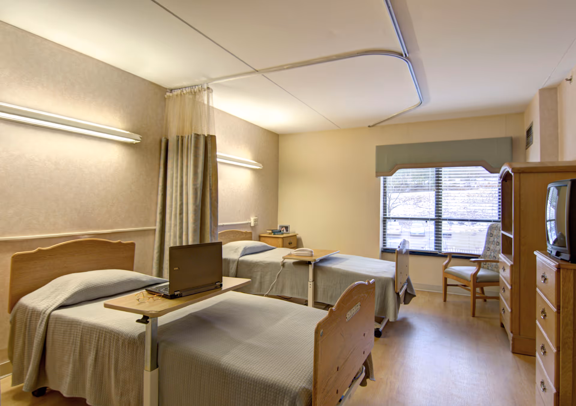 Two hospital-style beds with nightstands, laptop on a desk, and a window in a patient room.