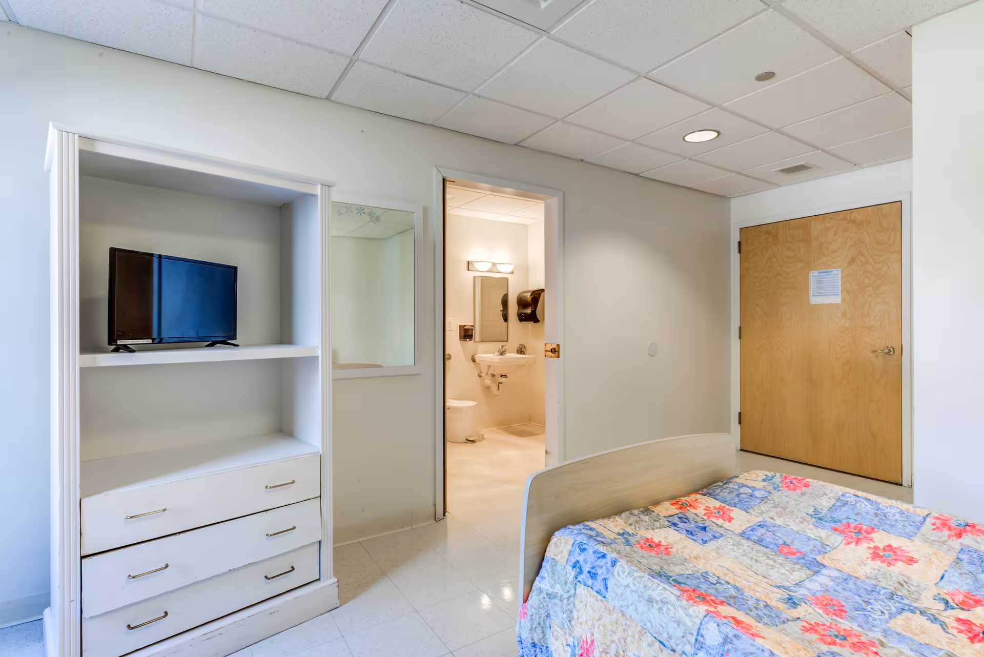 A senior living facility bedroom with a bed, quilt, dresser, TV, and adjacent bathroom doorway.