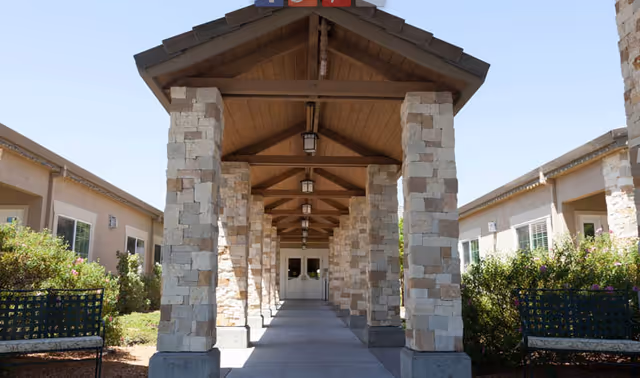 Covered stone-columned walkway leading to the entrance doors of a single-story senior living building with benches and landscaping on either side.