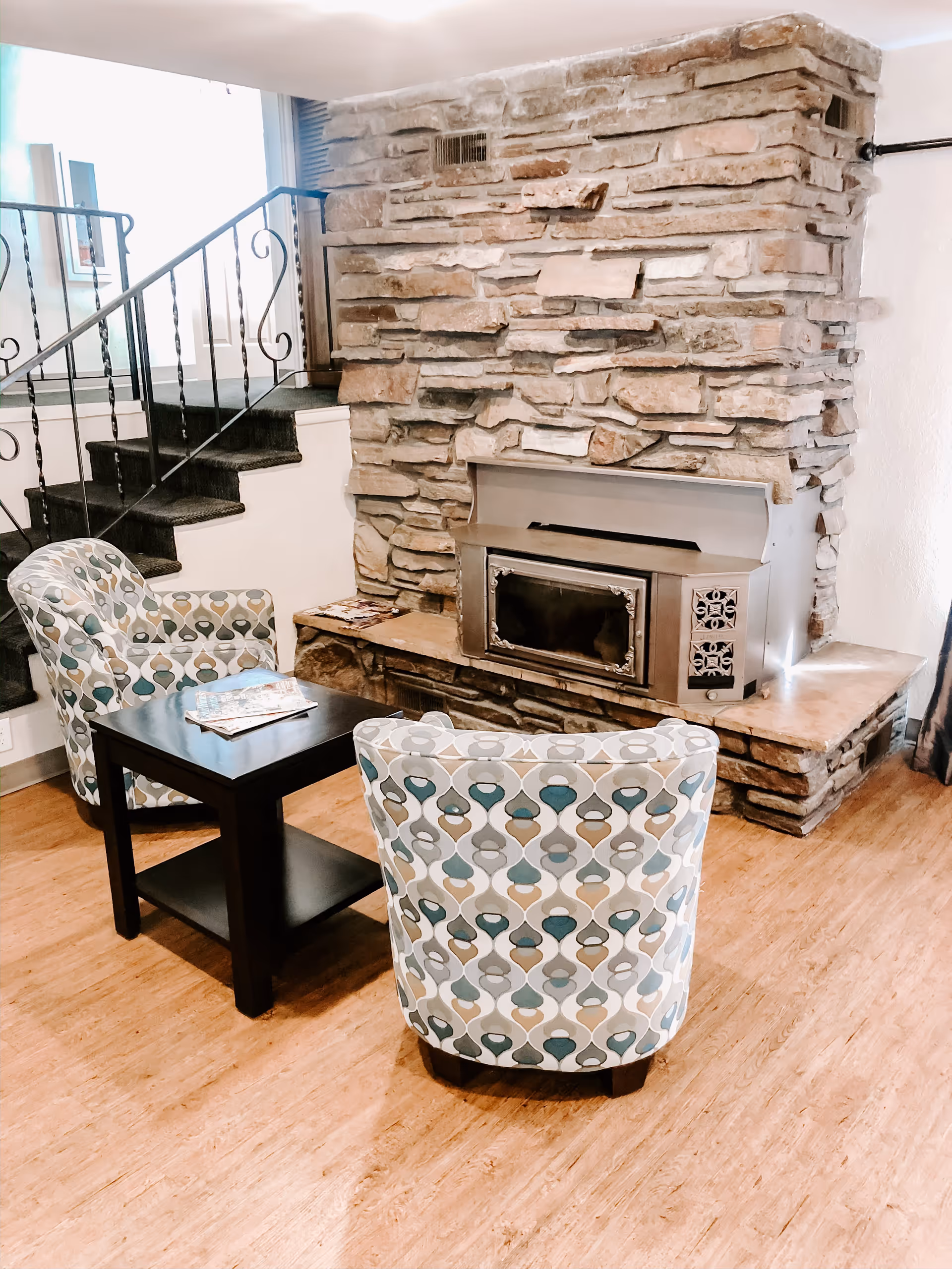 Two patterned armchairs and a small black table facing a stone fireplace with a staircase in the background.