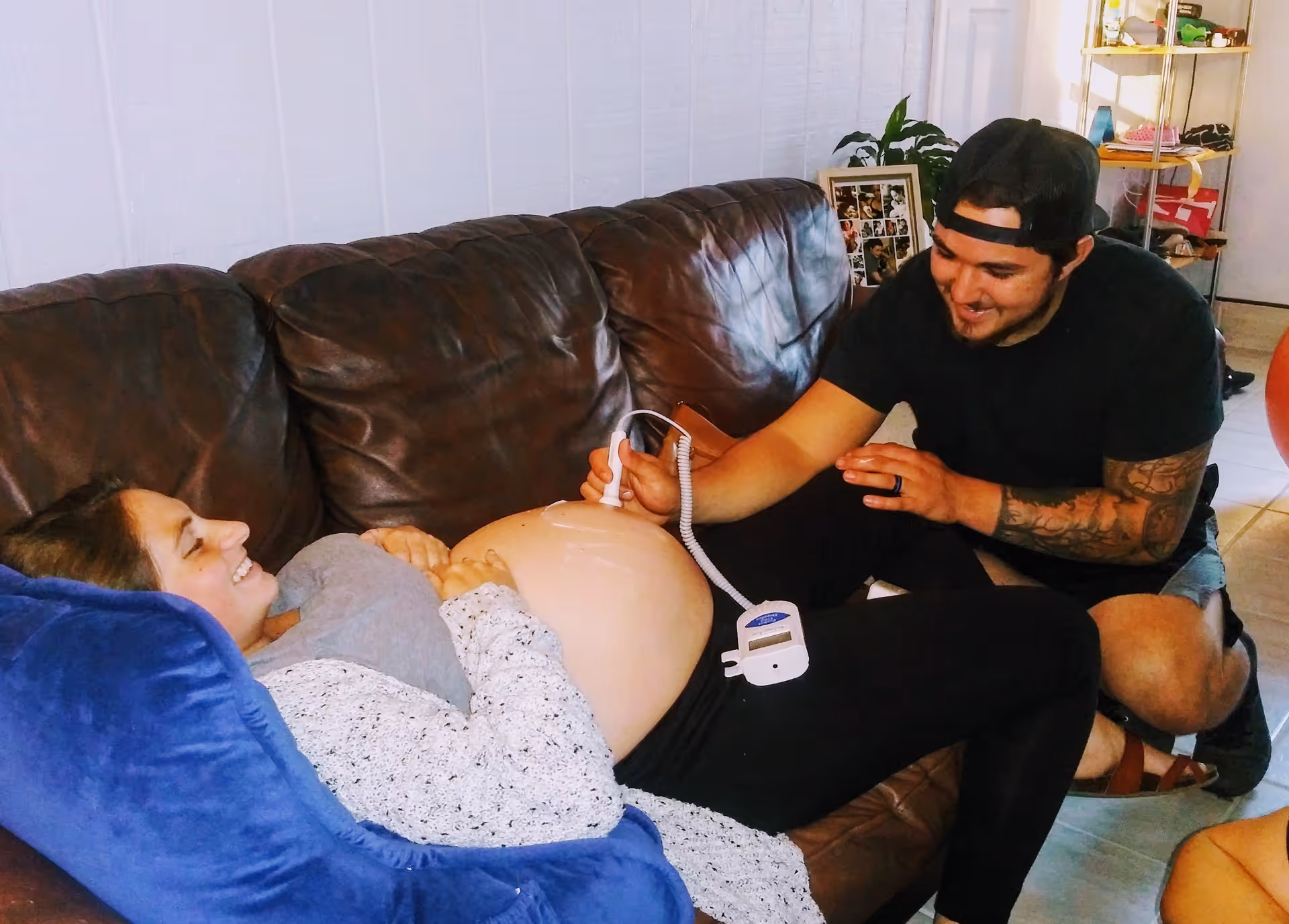 Pregnant woman on a couch with partner using a fetal monitor in living room.