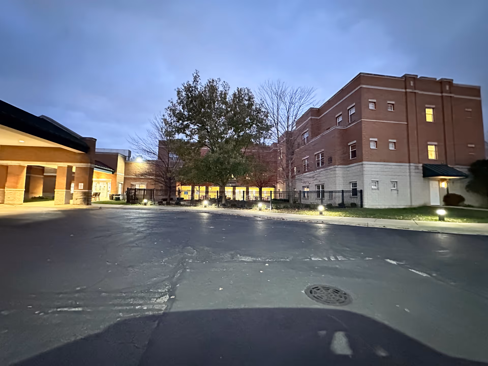 Exterior view of a multi-story brick building at dusk with illuminated windows and outdoor lighting. There is a paved driveway or parking area in the foreground, a tree in the center, and a covered entrance on the left side.