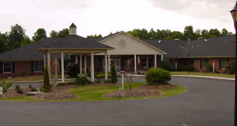 Exterior view of a single-story brick building with a covered entrance and a circular driveway, surrounded by landscaped greenery and trees in the background under a cloudy sky.