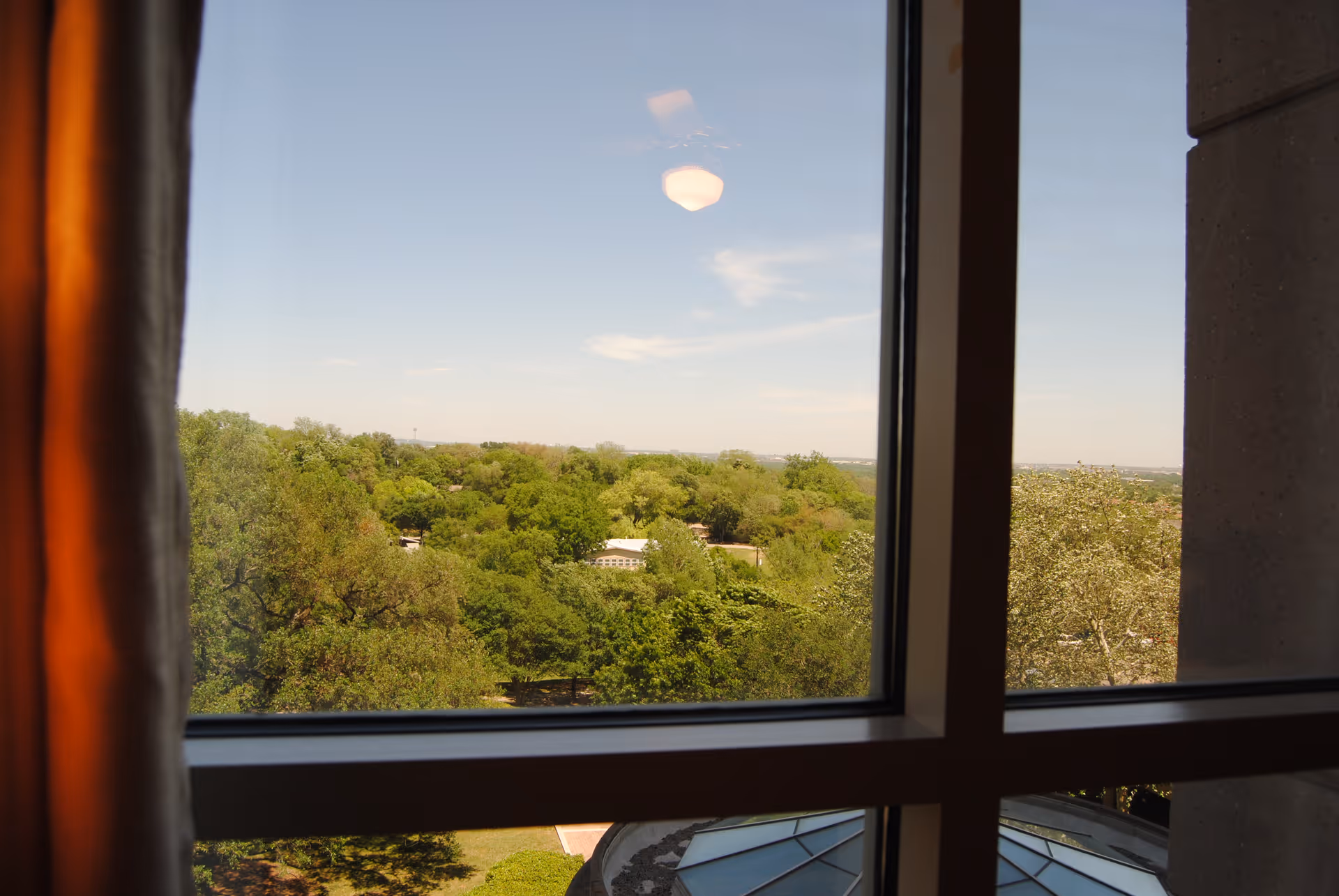 View through a window showing a landscape with many green trees under a clear blue sky. Part of a curtain and a building structure are visible on the sides of the window.