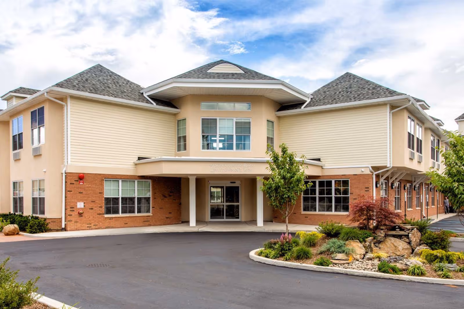 Front exterior view of a two-story senior living facility building with beige siding and brick accents, a covered entrance, several windows, and landscaped greenery including small trees and shrubs in front.