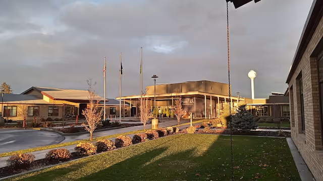 Front entrance of the Walla Walla Veterans Home with flagpoles, landscaped beds, and a water tower in the background at sunset.