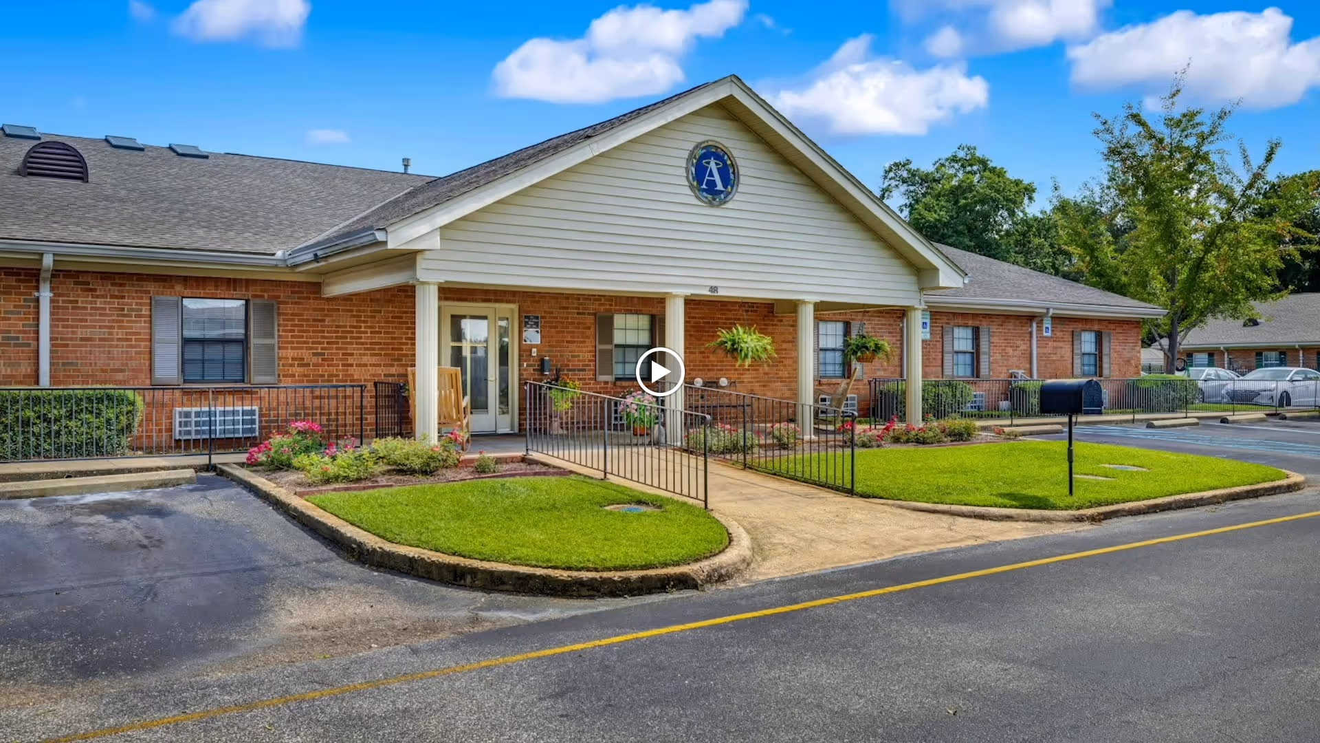 Exterior view of a single-story brick building with a covered entrance featuring a ramp and handrails. The building has a gable roof with a circular emblem displaying the letter 'A' above the entrance. There are well-maintained green lawns and flower beds on either side of the walkway leading to the door. Several windows with shutters are visible, along with a mailbox near the street. The sky is blue with some clouds.
