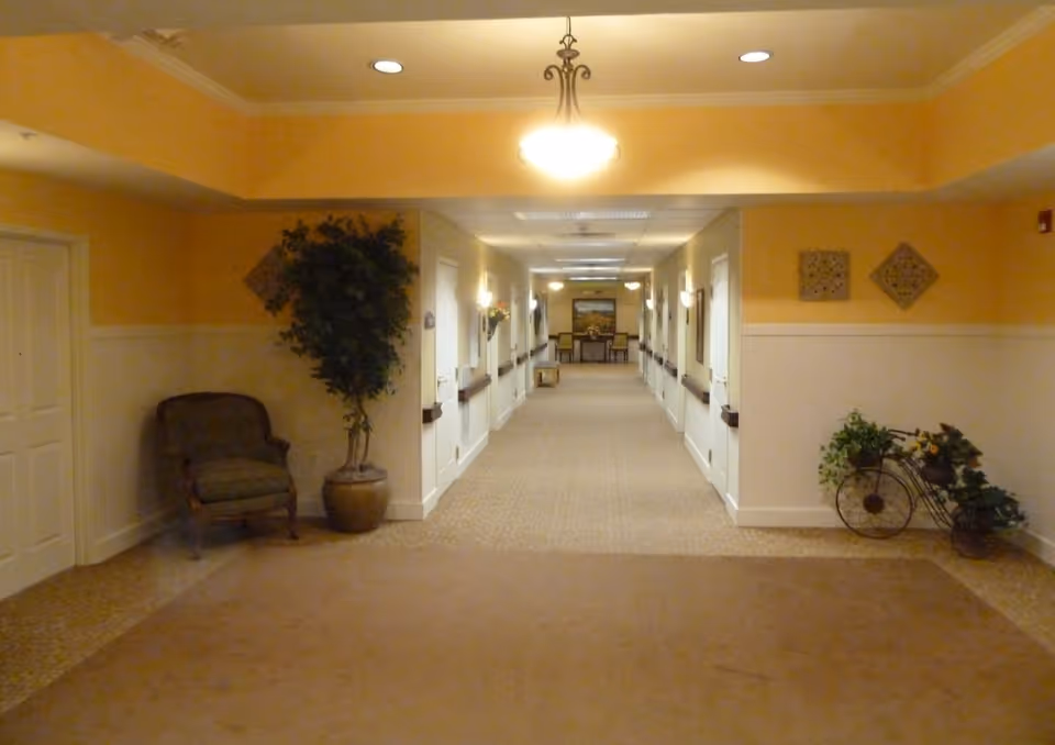 Well-lit interior hallway in a senior living facility with seating, potted plants, and decorative accents leading to a distant sitting area.