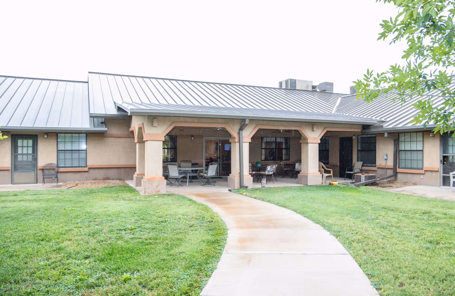 Front exterior of a single-story retirement community building with a covered patio, outdoor seating, and a walkway across a lawn.
