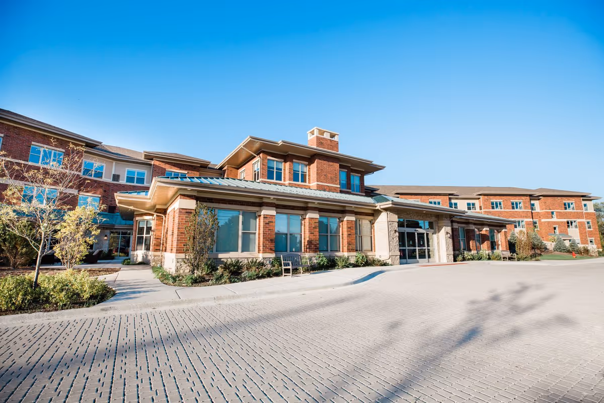Brick senior living facility building with a prominent entrance, large windows, landscaped beds, and a paved driveway under a clear blue sky.