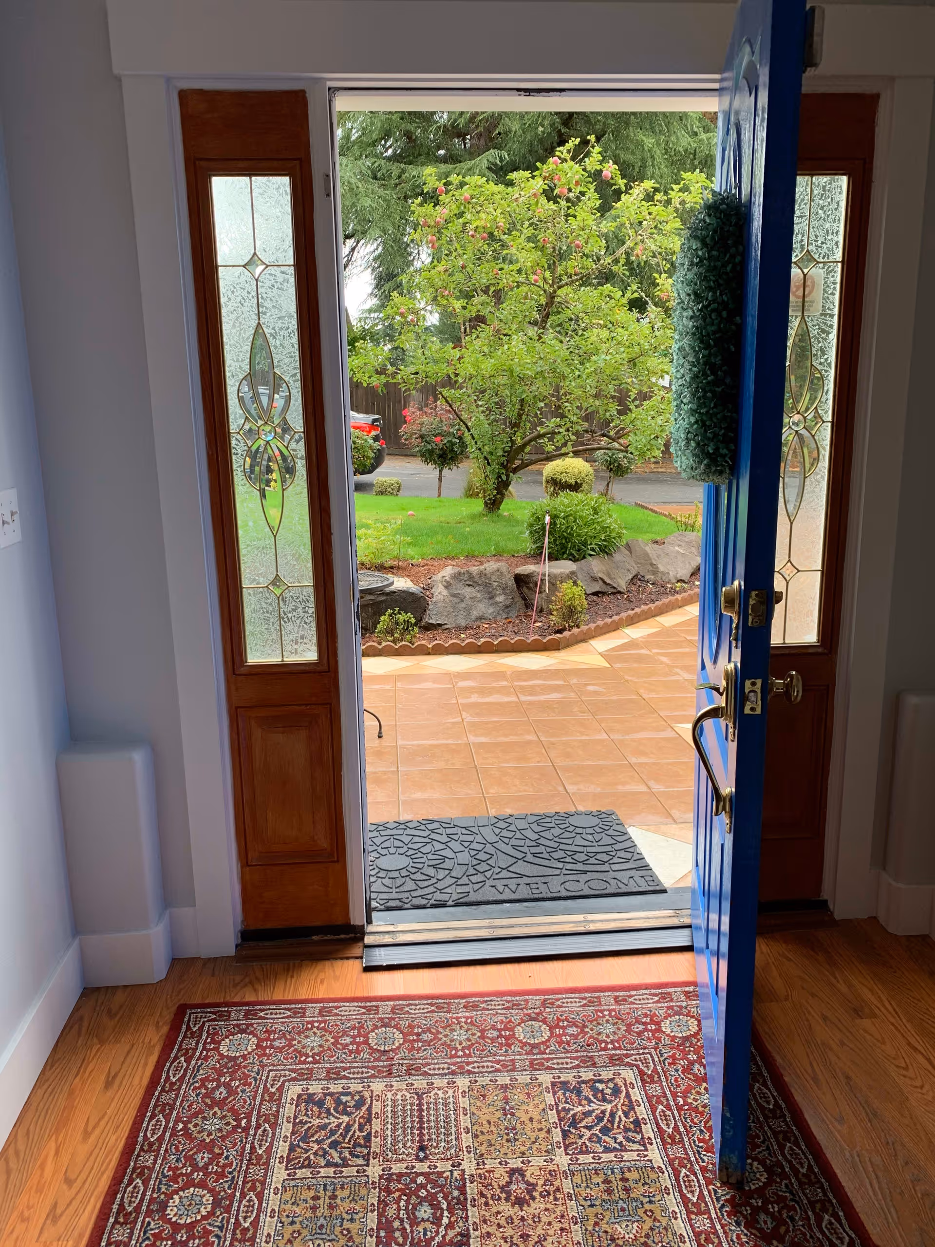 Open blue front door with decorative glass sidelights revealing a tiled porch, a welcome mat, and a landscaped yard with a tree.