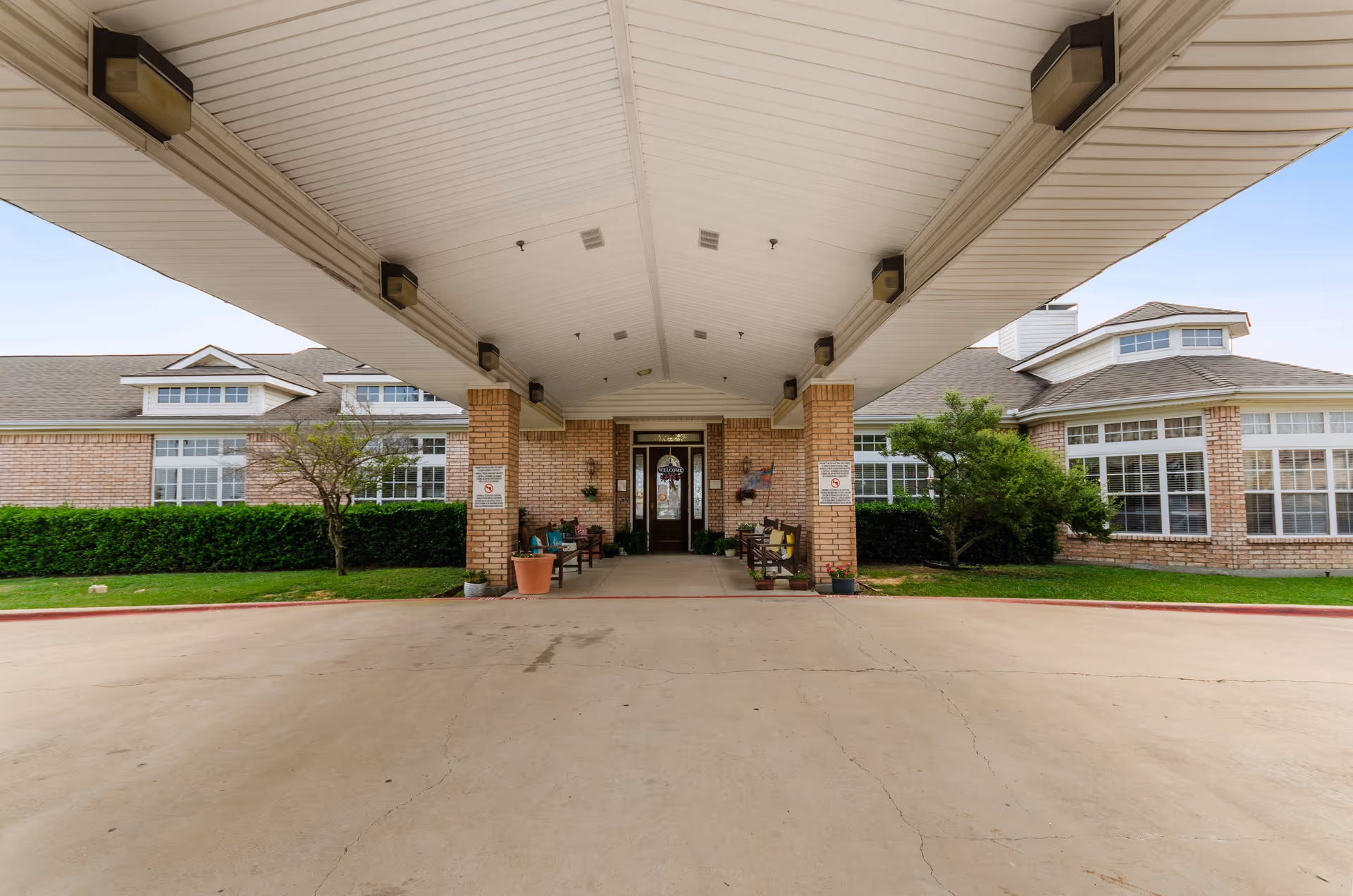 Covered porte-cochere entrance leading to the front doors of a brick assisted living building with benches and potted plants.