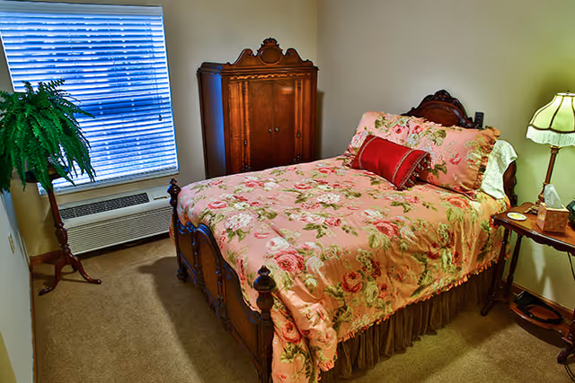 A cozy bedroom with a wooden bed frame and a floral pink bedspread with matching pillows. There is a wooden armoire against the wall, a small wooden side table with a lamp, and a green potted plant on a stand near a window with closed blinds.