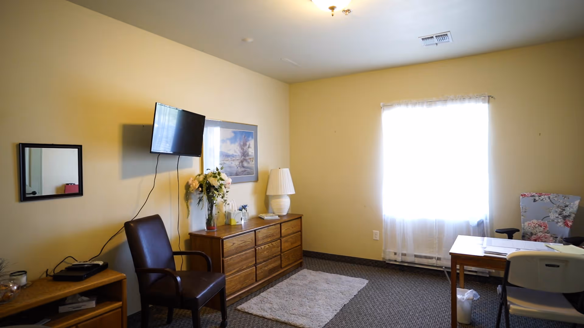 Senior living room with wall-mounted TV, dresser, chair, and bright window.